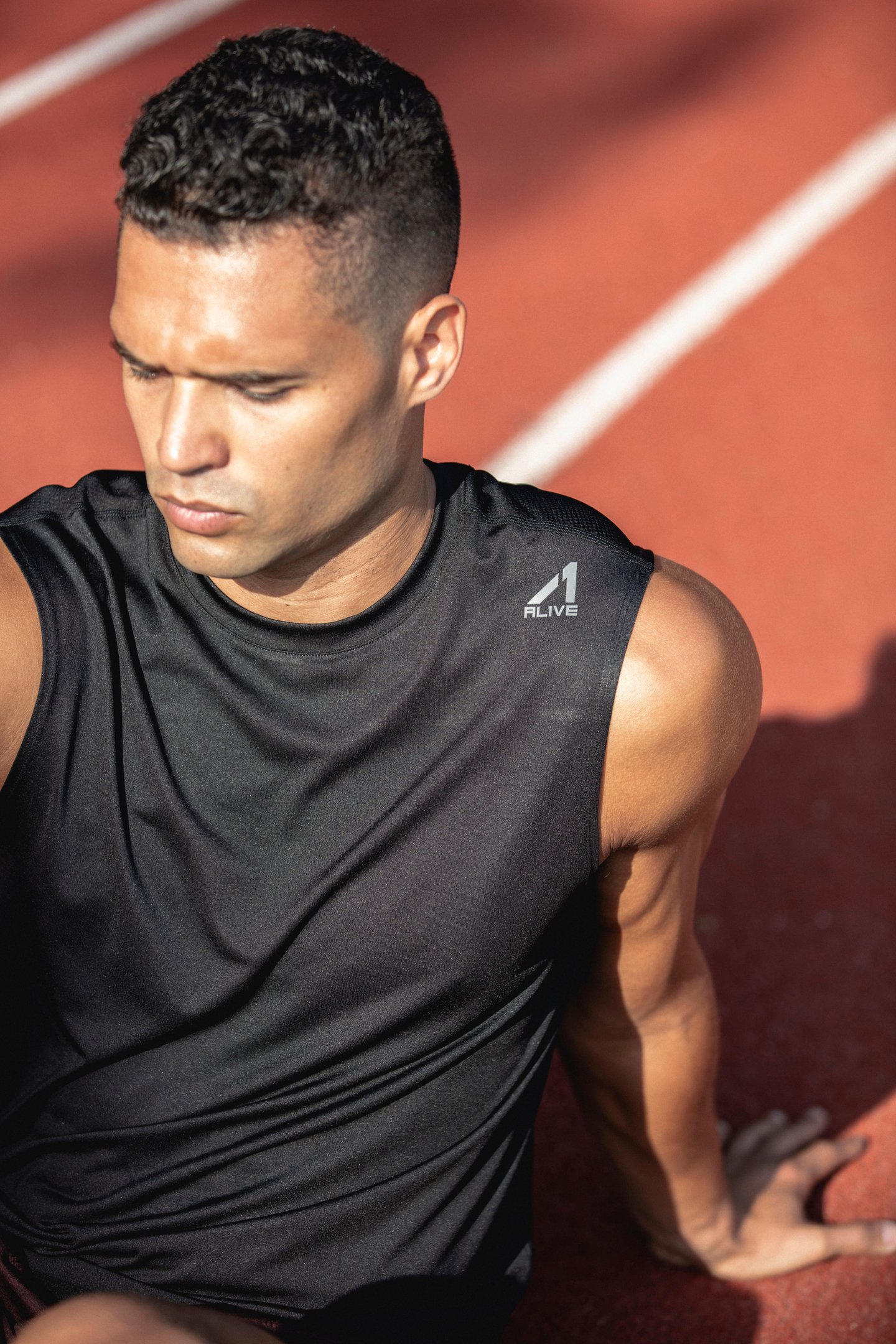 A close-up photo of a male athlete performing a seated stretch on a track field in the sun. He is wearing a black sleeveless top from Alive and has a focused look on his face.